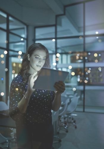 woman with a dark blouse and black slacks standing in an office conference room holding and looking at a computer tablet.